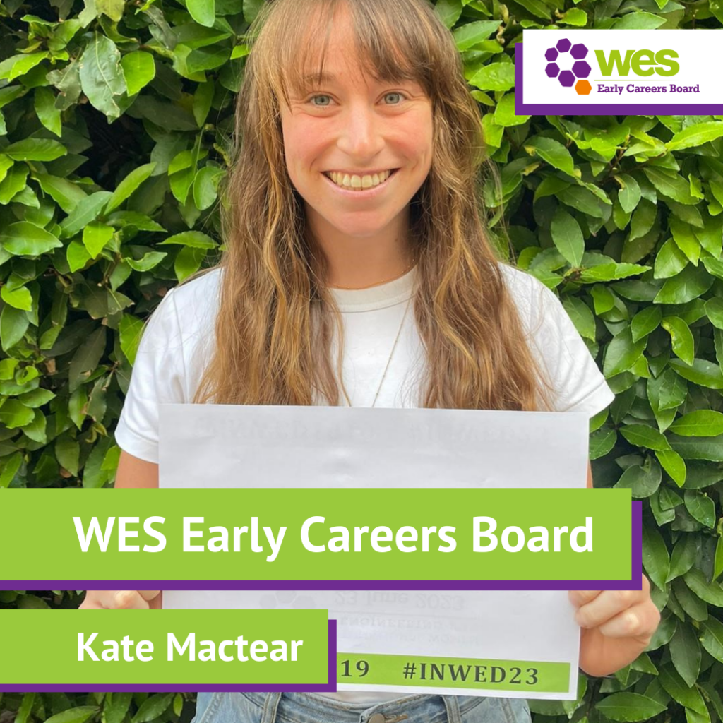 A young woman with long, wavy light brown hair and a white t-shirt, smiling and holding a sign that reads "WES Early Careers Board Kate Mactear" in front of a lush green leafy background. The top right corner has a purple and orange logo with the text "WES Early Careers Board."