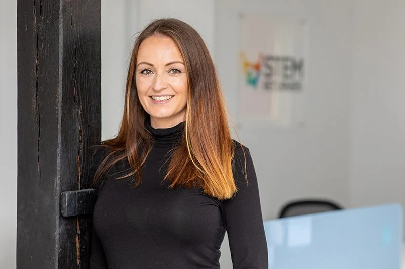 A smiling woman with long brown hair in a black turtleneck standing next to a dark wooden wall in an office setting.
