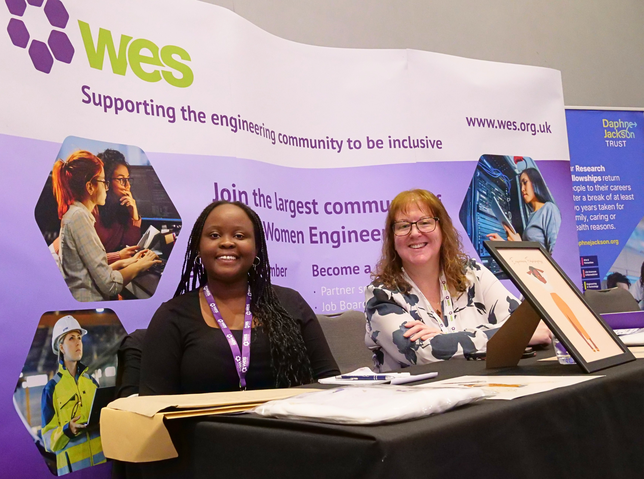 Two women sitting at a table with promotional materials at an event supporting women in engineering, with posters and banners in the background.