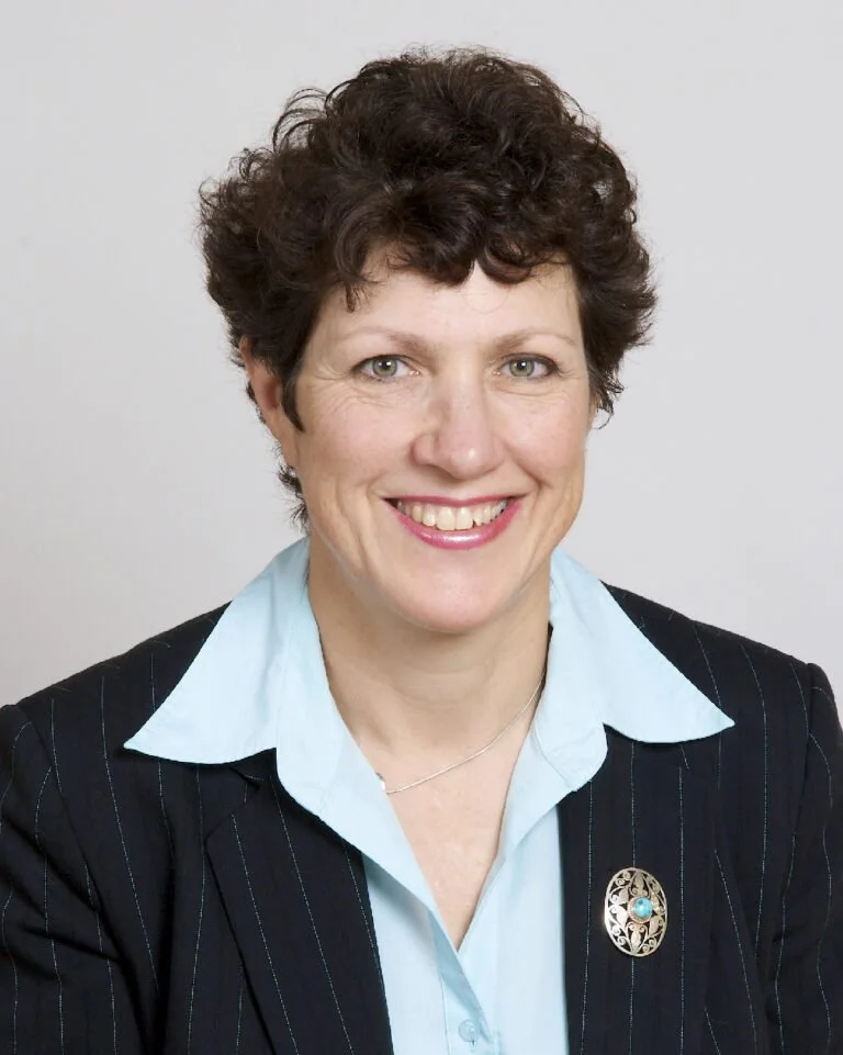 A woman with short curly brown hair smiling, wearing a black pinstripe blazer over a light blue shirt, with a decorative brooch on the blazer.