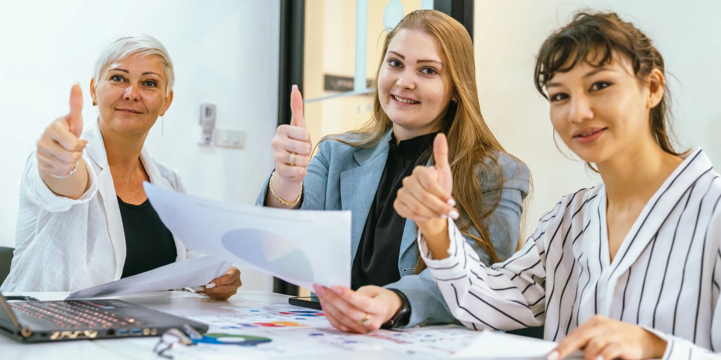 Three women sitting at a table, giving thumbs-up and smiling, with papers and a laptop in front of them.