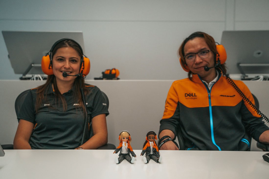Two people wearing headsets sitting at a table with two dolls between them, all in a professional setting.
