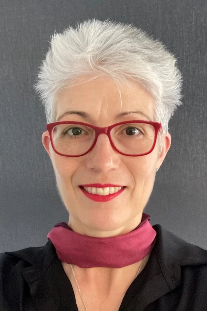 A smiling woman with short gray hair, red glasses, red lipstick, wearing a black top, a purple neck scarf, and a delicate necklace, standing against a gray background.