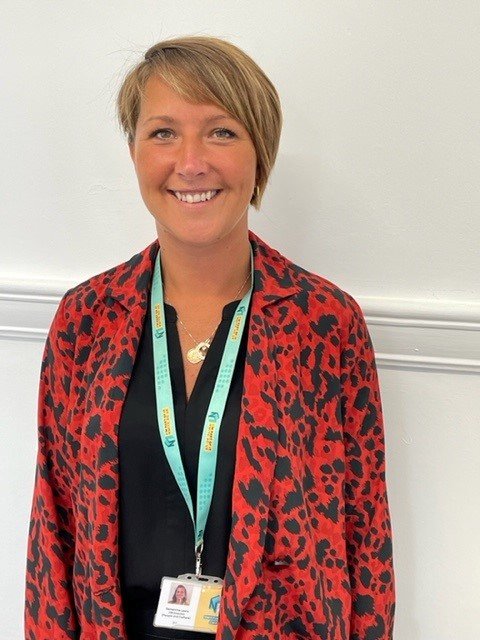 A woman with short blonde hair smiling, wearing a red and black leopard print blazer, a black top, and a lanyard with an ID badge.