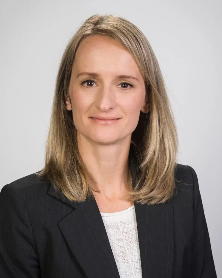 A professional woman with shoulder-length blonde hair, wearing a black blazer over a white top, smiling softly against a plain gray background.