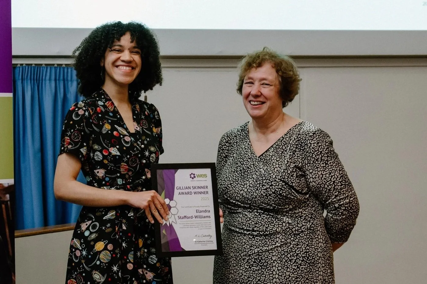 A woman receiving an award certificate from another woman in a room with a white wall and blue curtains. Both women are smiling.