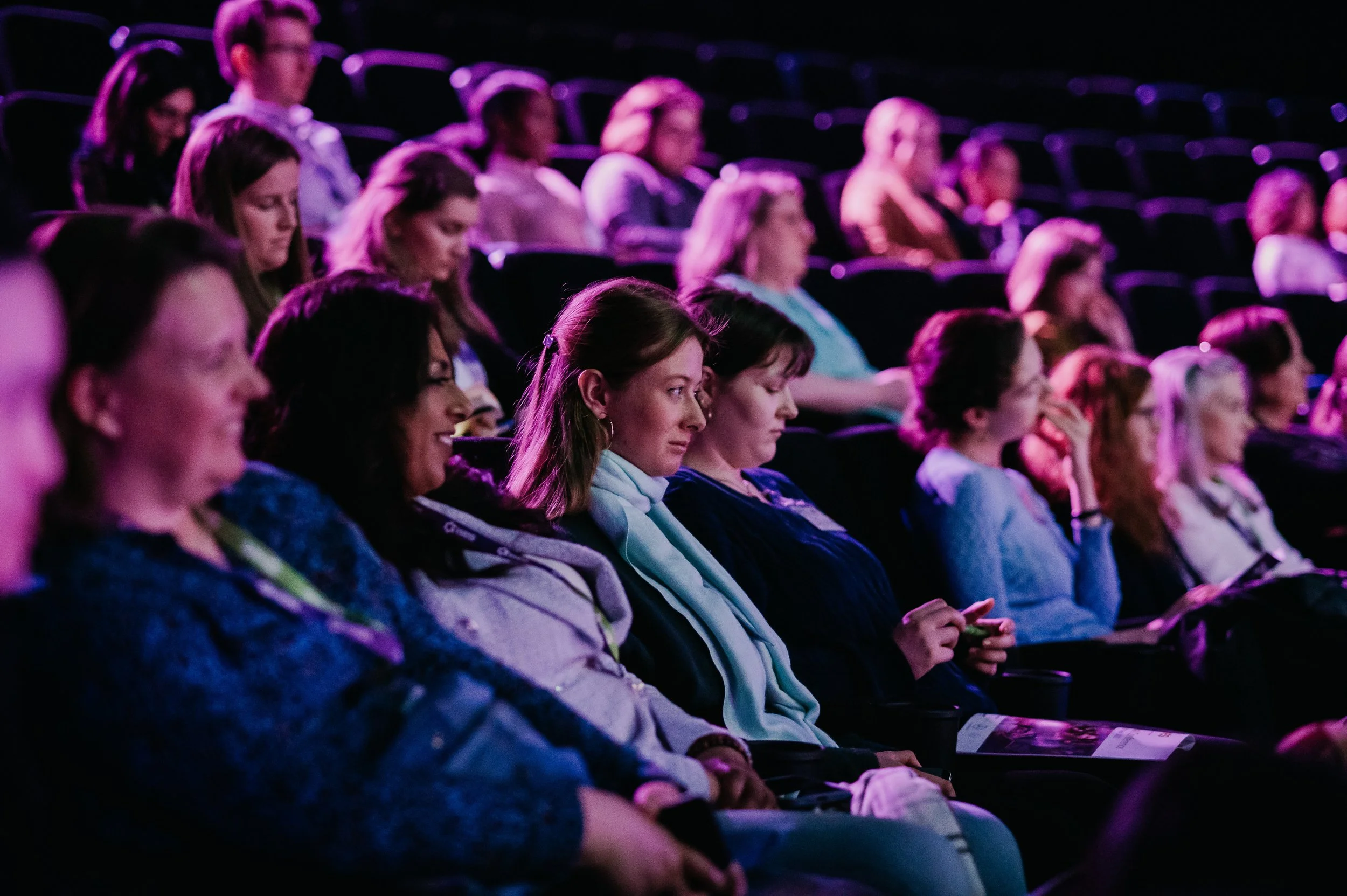 Audience seated in a dark theater or auditorium, illuminated by purple lighting, with some people looking down or at their phones.