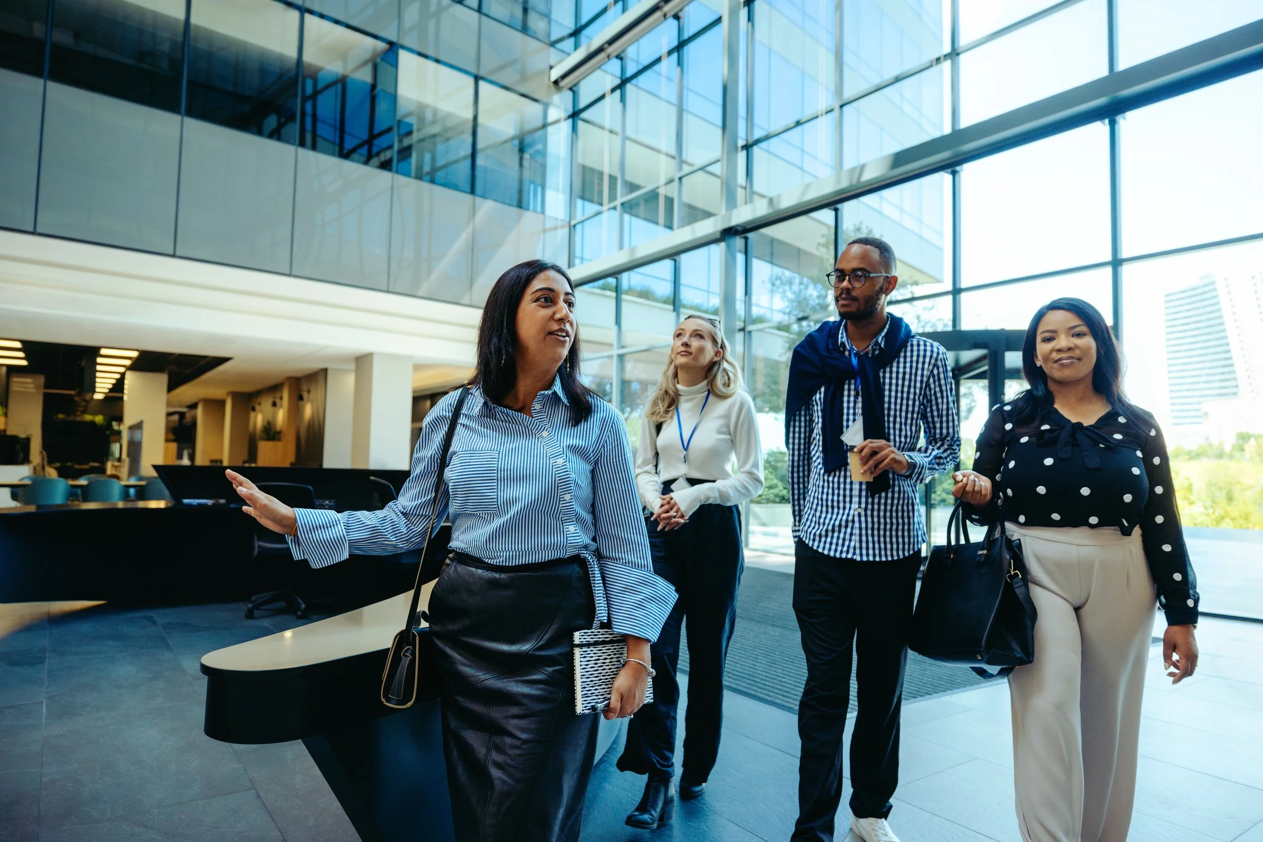 Four diverse people in business casual attire walking and conversing in a modern, glass-walled office lobby with natural light and contemporary architecture.
