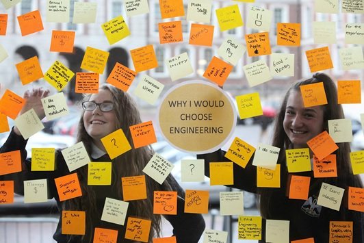 Two smiling women are looking at a glass wall covered with colorful sticky notes. In the center, there is a large white circle with the words 'WHY I WOULD CHOOSE ENGINEERING' written inside.