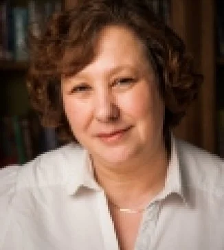 A woman with curly brown hair wearing a white shirt, smiling softly, standing in front of a bookshelf.