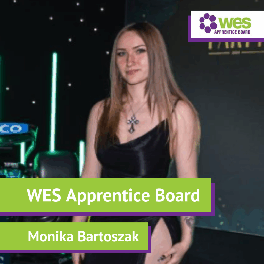 A woman with long brown hair wearing a black dress and a cross necklace standing against a dark background with green and purple lighting. Text overlays read "WES Apprentice Board" and "Monika Bartoszak." A logo for WES Apprentice Board is in the top right corner.