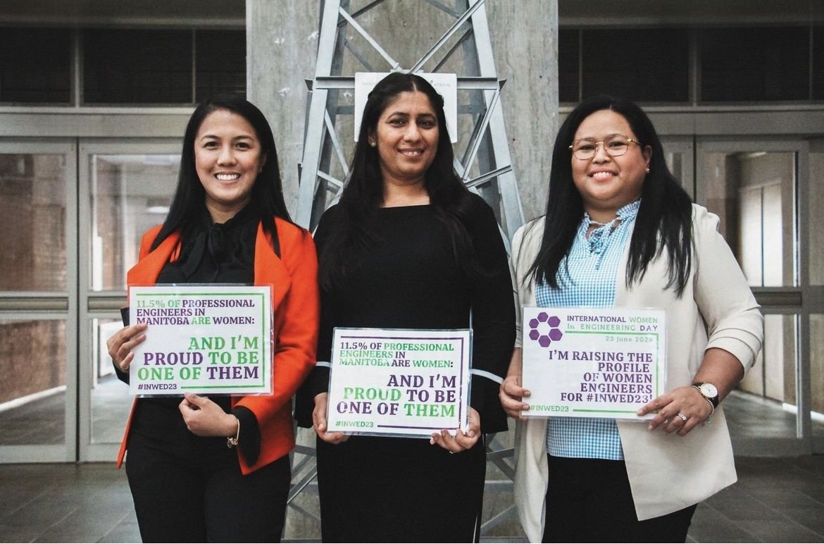 Three women standing indoors, holding signs during International Women in Engineering Day 2023. The woman on the left is wearing a red blazer and black top, the woman in the middle is dressed in black, and the woman on the right is wearing glasses, a beige blazer, and a light blue checkered shirt.