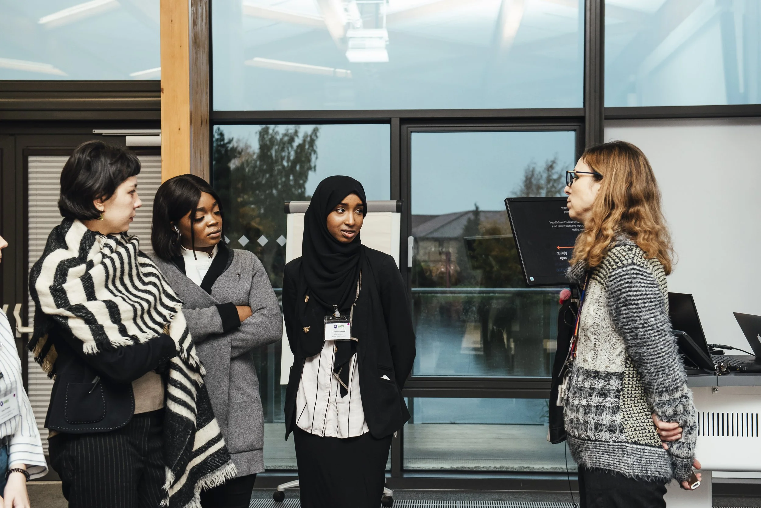 A group of diverse women in a professional setting, engaging in a discussion with one woman speaking to the others. The background features large windows and a modern office environment.
