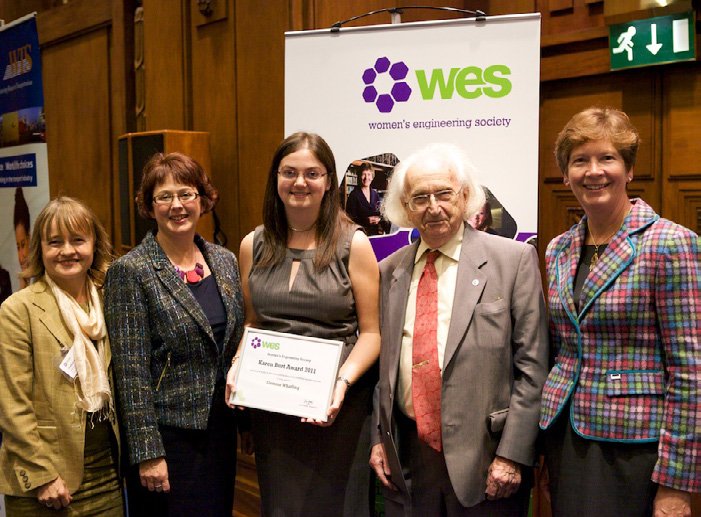 Five women and one elderly man standing together at a Women's Engineering Society event, with a banner in the background labeled 'WES' and 'women's engineering society'. One woman in the center is holding a certificate.