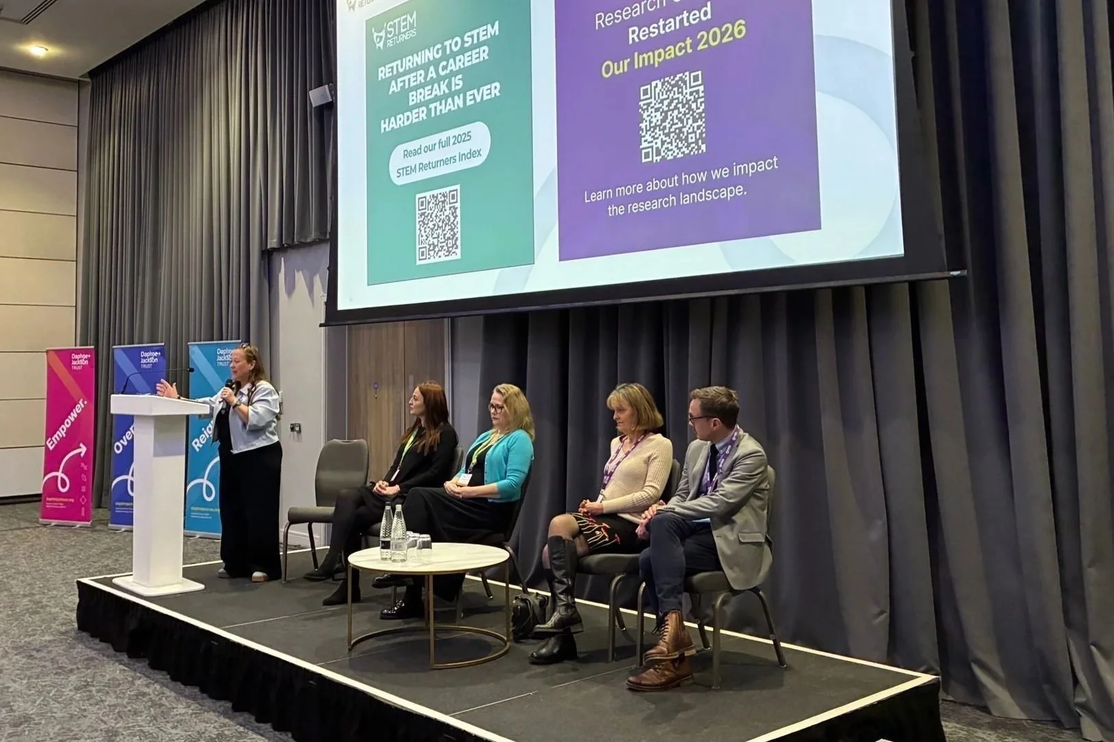 A woman is speaking at a podium during a panel discussion at a conference. Five people are seated on stage, and a large screen behind them displays a presentation slide with QR codes and text. There are vertical banners with the words "Empower" and other keywords in the background.