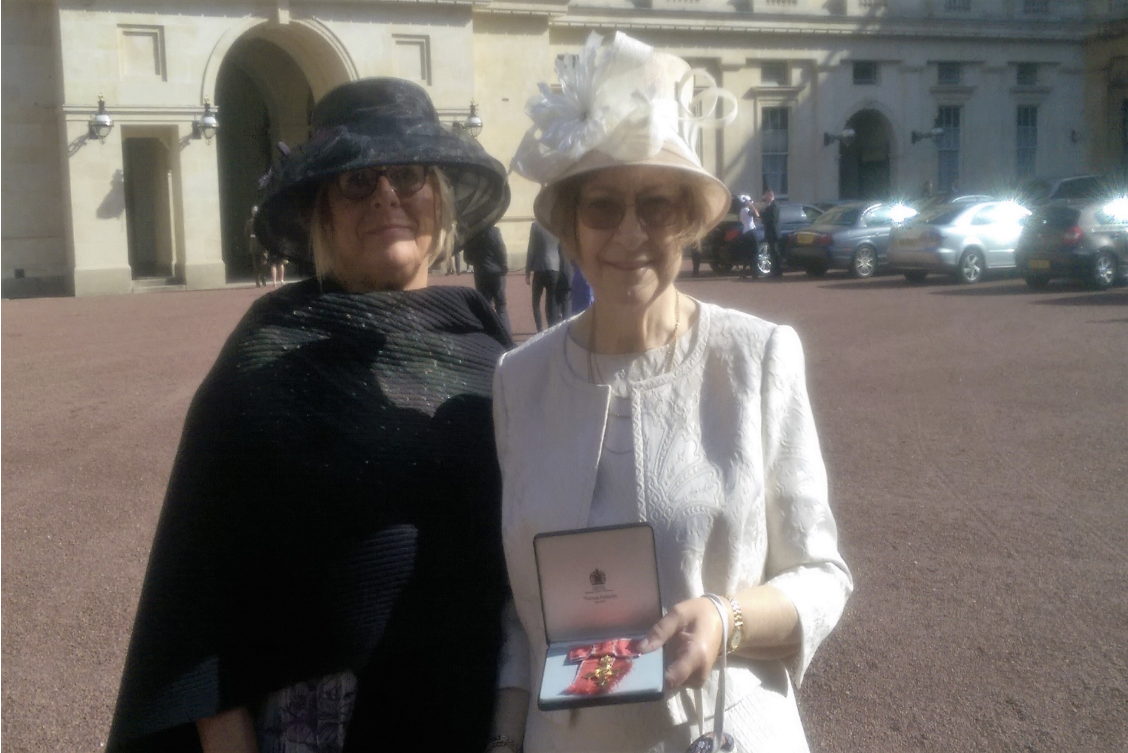 Two women standing outdoors in front of a historic building with parked cars in the background. One woman is wearing a large black hat and sunglasses, and the other is wearing a large white hat with a flower decoration and sunglasses. The woman in white is holding a framed certificate or award.