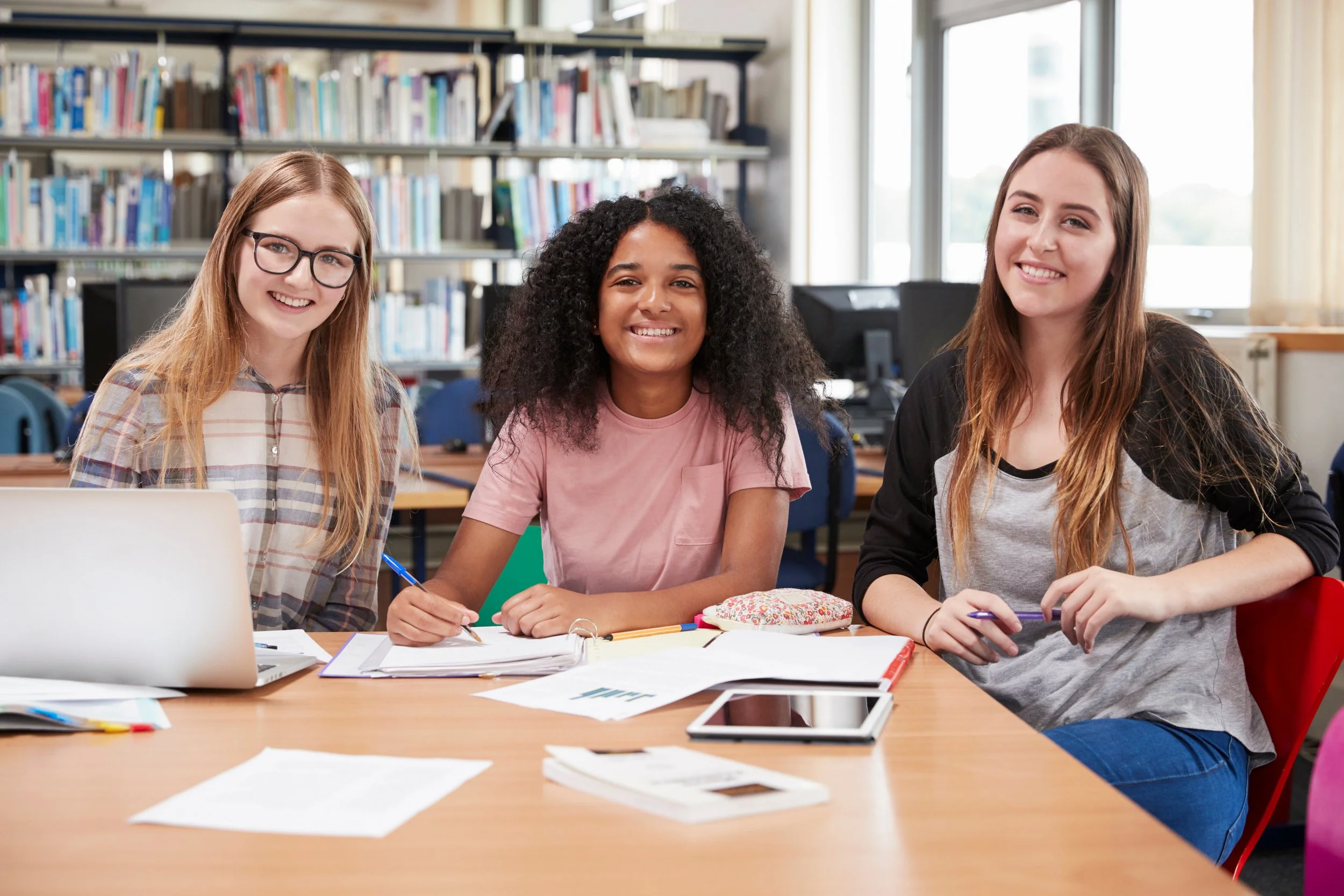Three young women sitting at a table in a library, smiling at the camera, with bookshelves and windows in the background.