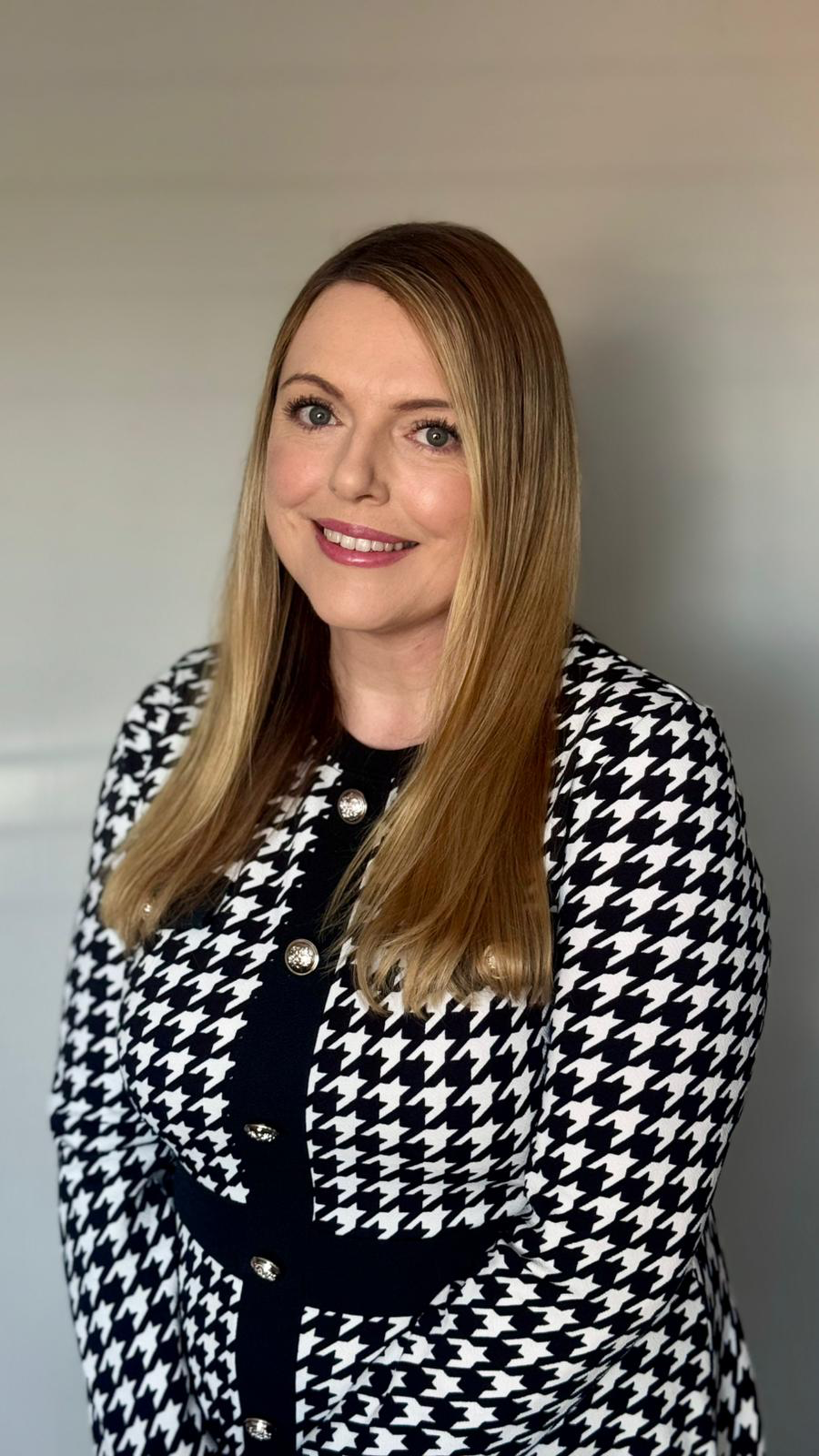 A woman with straight, shoulder-length blonde hair, blue eyes, and light skin, smiling at the camera, wearing a black and white houndstooth patterned dress with decorative buttons.