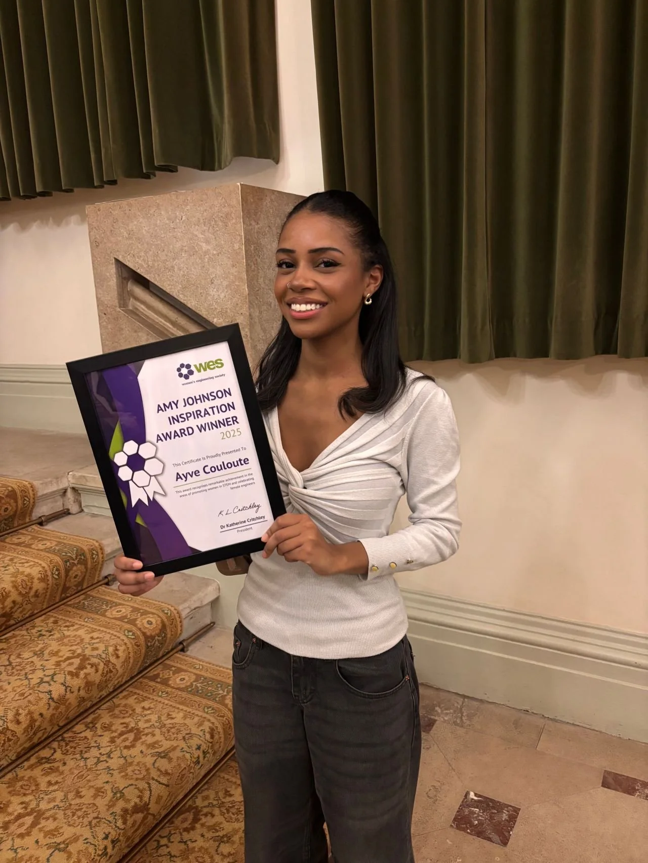 A woman smiling and holding a framed award certificate in a room with green curtains and patterned seating. The certificate recognizes her as the Amy Johnson Inspiration Award Winner 2025 from WES.