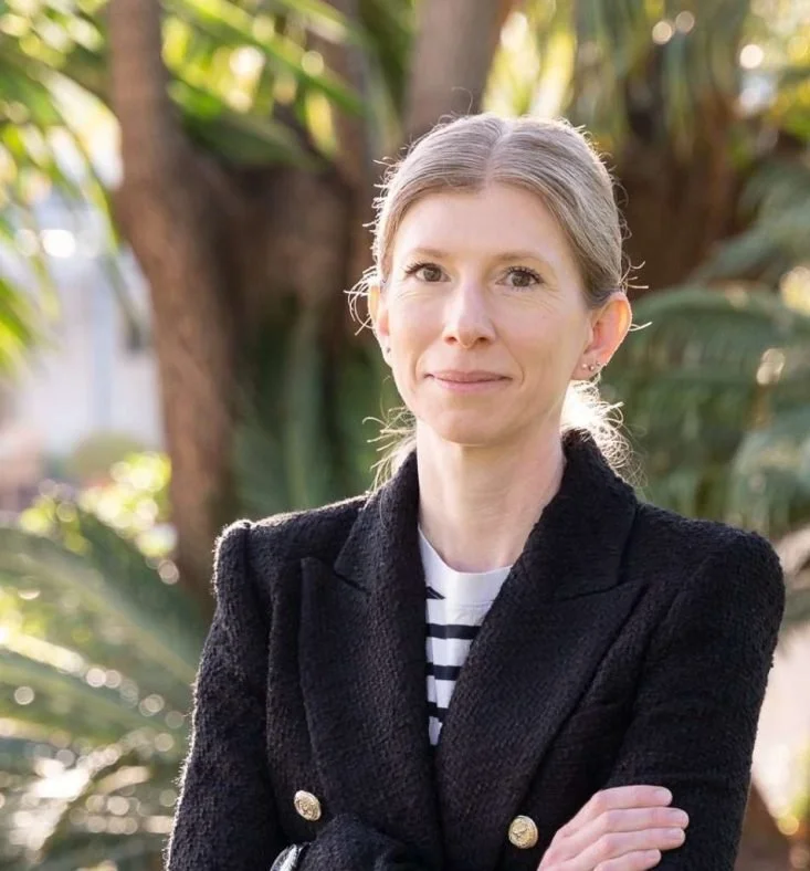 A woman with gray hair in a ponytail, wearing a black blazer with gold buttons over a striped shirt, standing outdoors in front of trees and greenery.