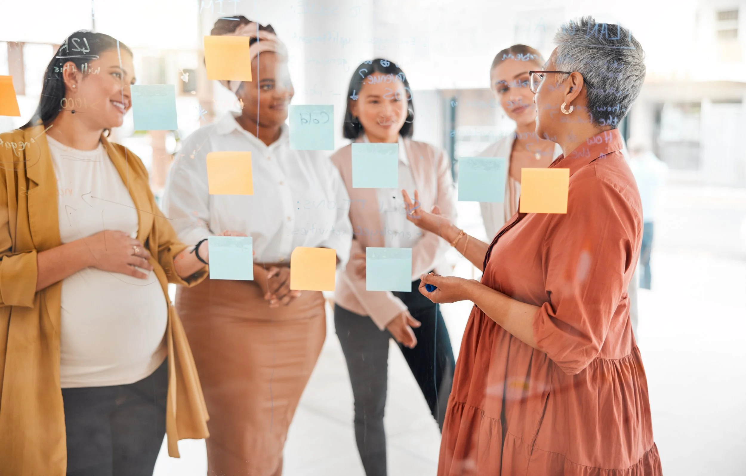 A diverse group of five women having a discussion behind a glass wall with colorful sticky notes, in a bright modern office.