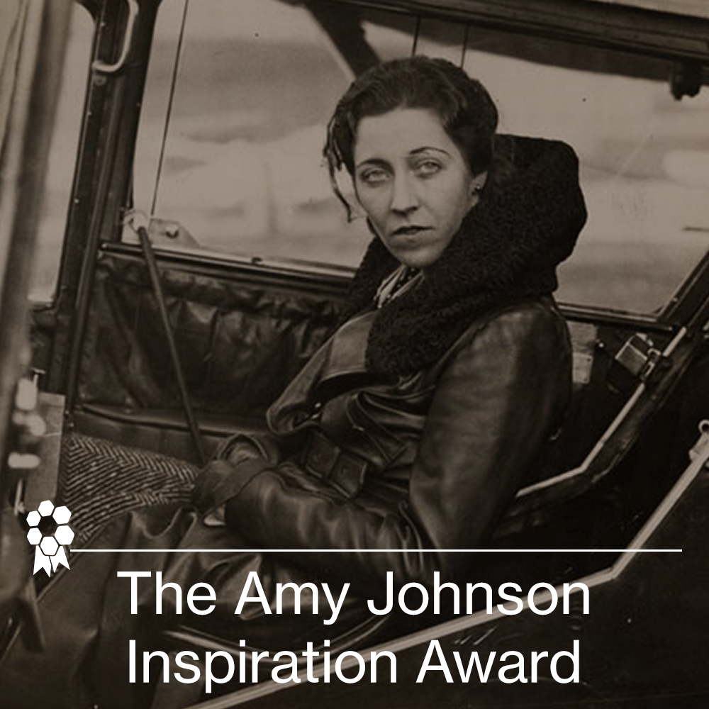 A woman with dark, wavy hair sitting in the driver's seat of a vintage car, looking serious, wearing a dark leather jacket with a scarf. The photo is in black and white.