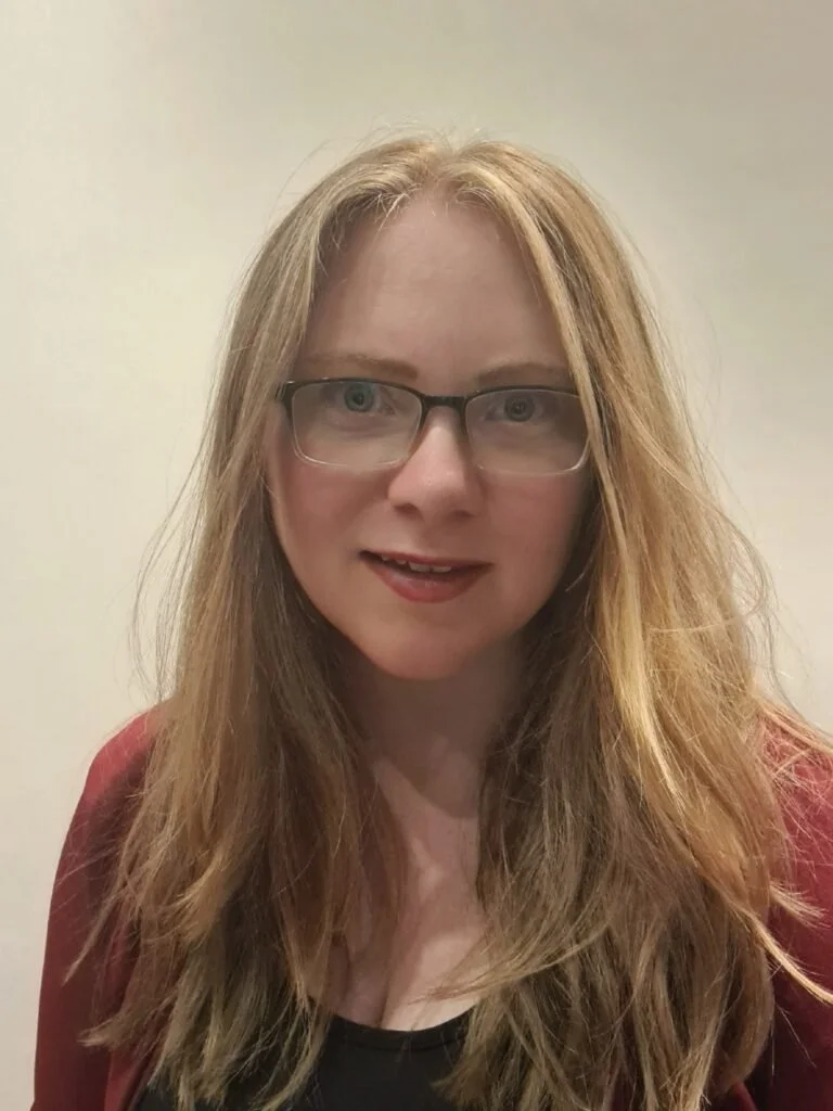 A woman with long, wavy, red hair and glasses looking at the camera with a slight smile, wearing a dark top and a reddish jacket, standing against a plain beige wall.