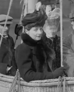 A woman wearing early 20th-century clothing and a hat standing in a hot air balloon basket, surrounded by other people.