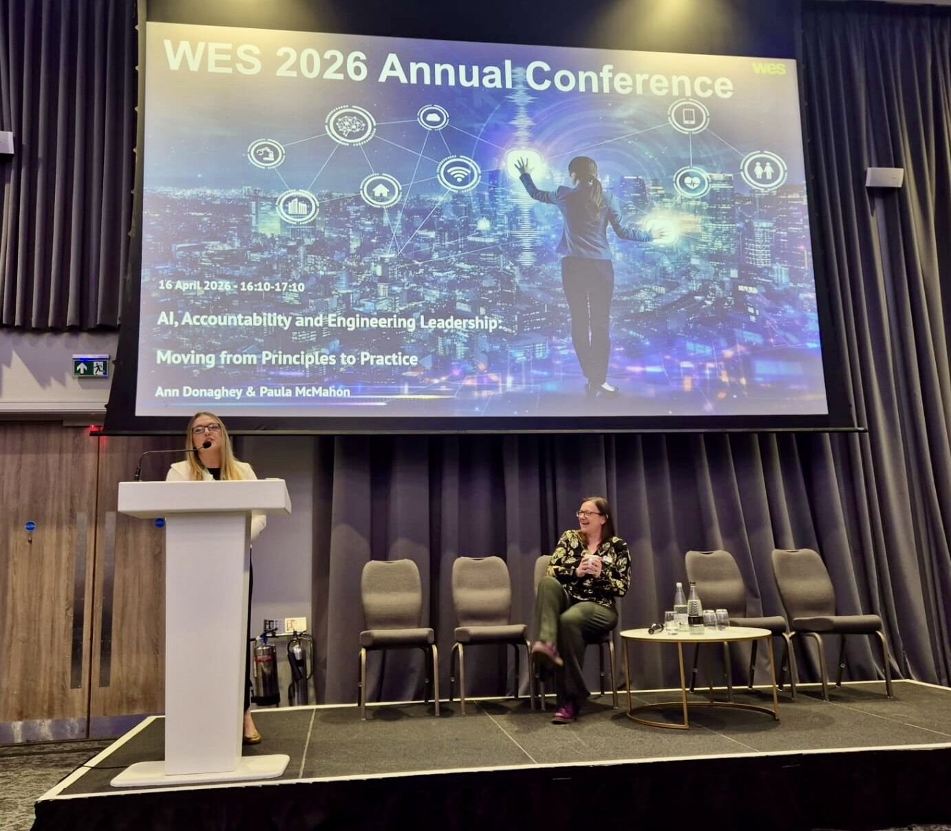 Conference room with a large screen displaying the title 'WES 2026 Annual Conference,' and two women speaking and sitting on stage. The screen shows a cityscape with interconnected icons representing technology and innovation.