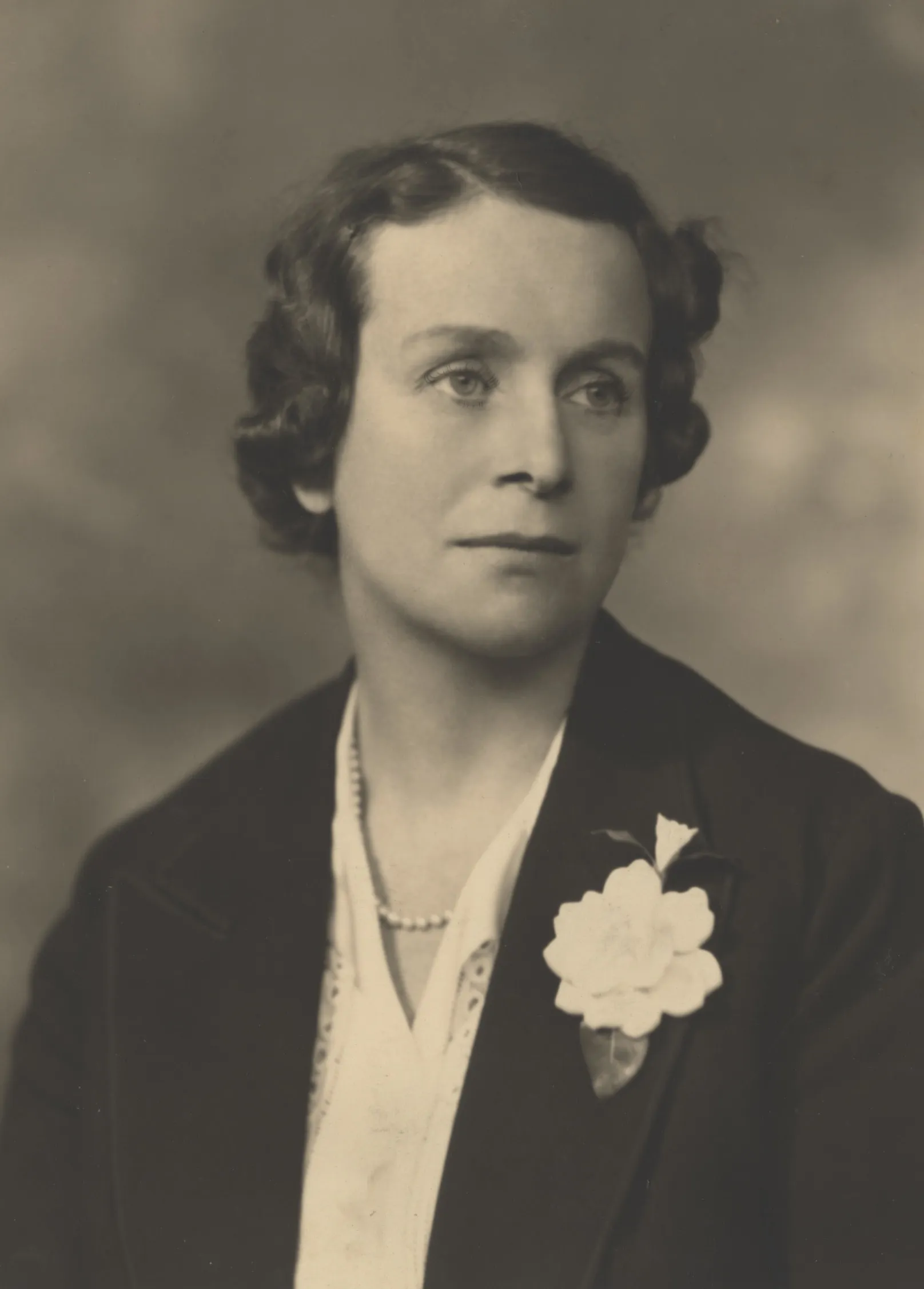 Black and white photograph of a woman with short, curly hair, wearing a dark blazer, a light-colored blouse, a string of pearls, and a large white flower pinned to her blazer.
