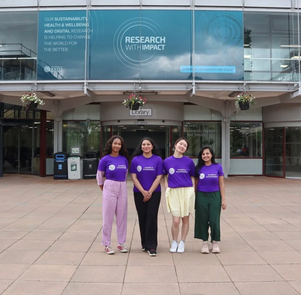 Four women in purple shirts standing outside a university building, smiling. The building has a sign for a library and banners promoting research impact.