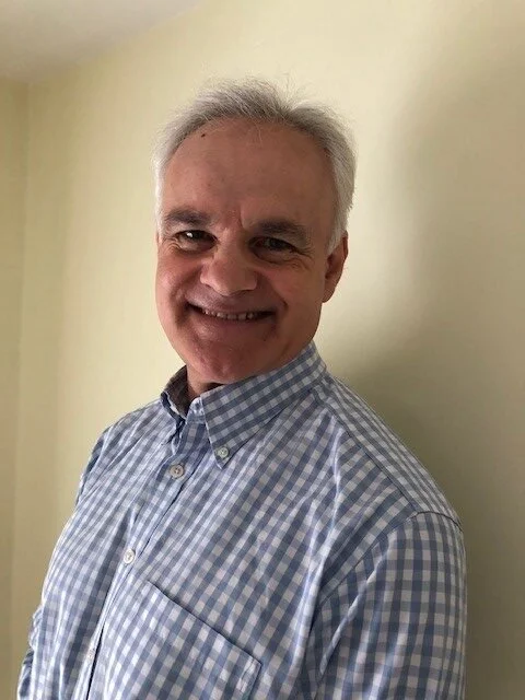 A middle-aged man with gray hair smiling, wearing a checkered button-down shirt, standing against a plain light-colored wall.