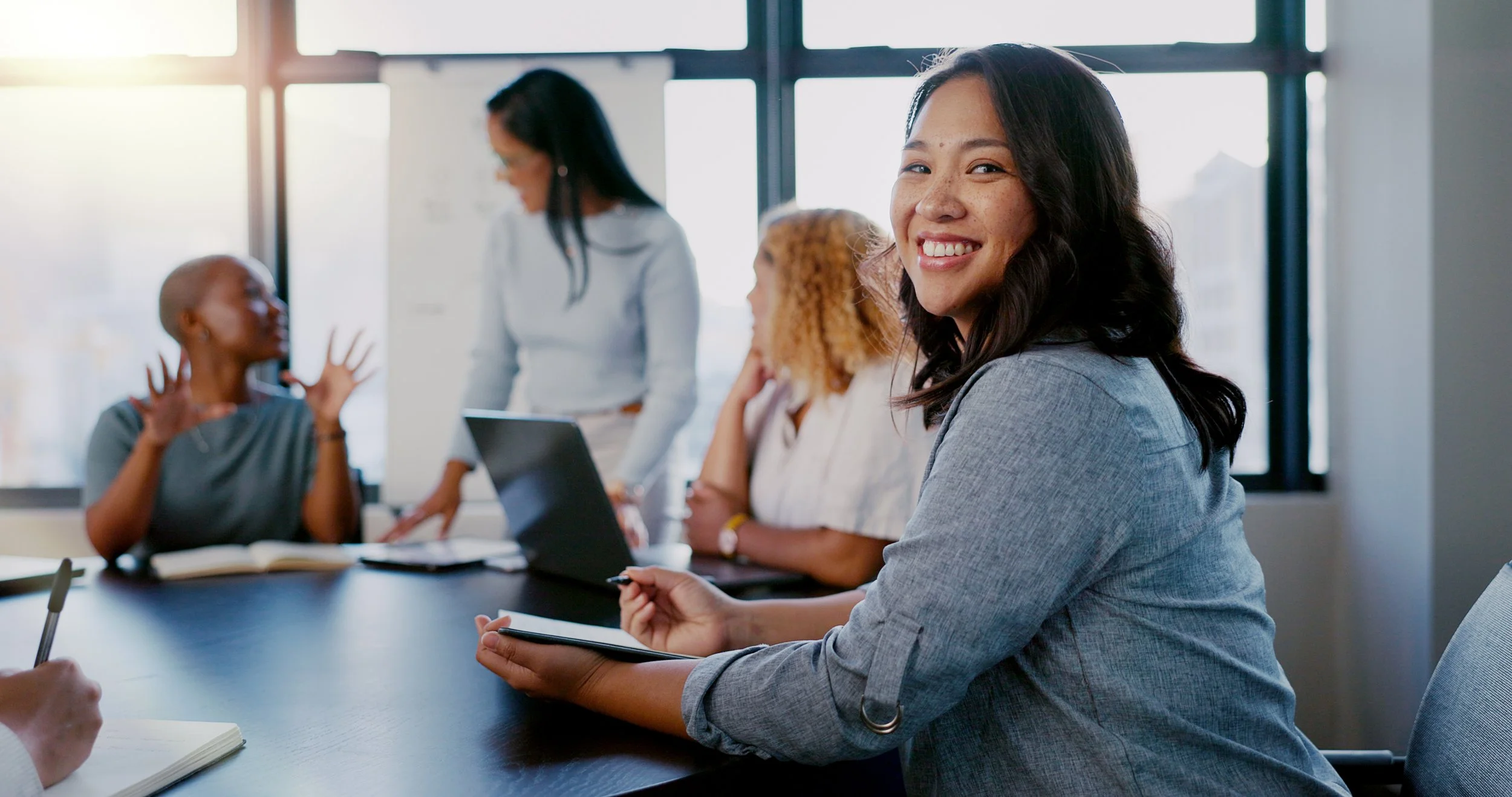 Young woman smiling during a meeting with colleagues in a modern office, with large windows, a laptop, and notebooks on the table.