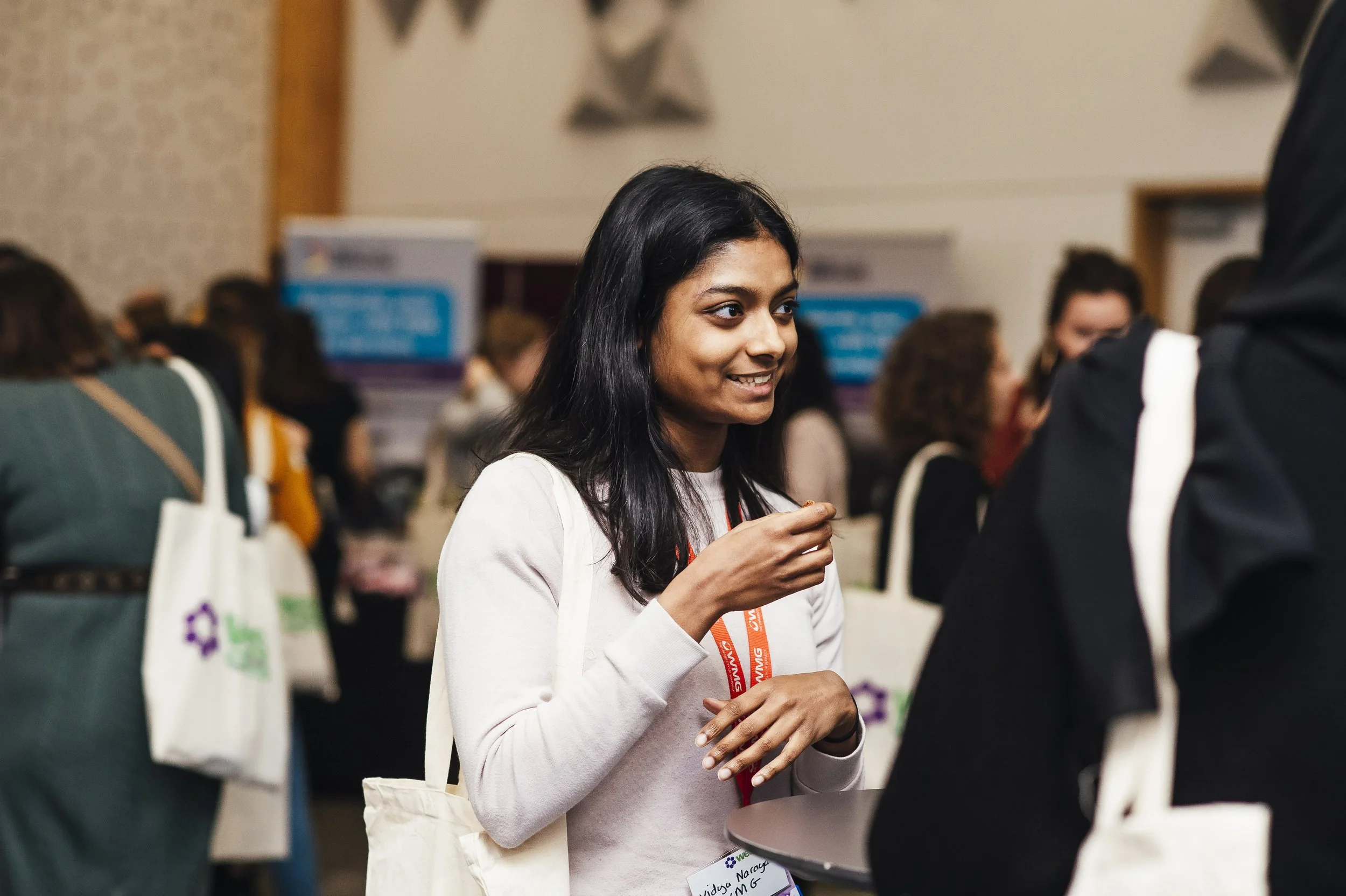 A young woman with black hair and medium skin tone smiling and engaging in conversation at a conference or event, wearing a white top, with a red lanyard around her neck, surrounded by other attendees.