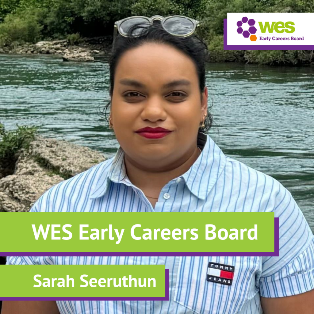 A person standing outdoors by a river with trees in the background, wearing a striped shirt and sunglasses on their head, and a logo for WES Early Careers Board and a name tag reading Sarah Seeruthun.
