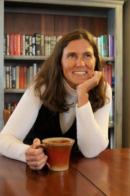 A woman with brown hair sitting at a wooden table, holding a coffee mug, in front of a bookshelf filled with colorful books.