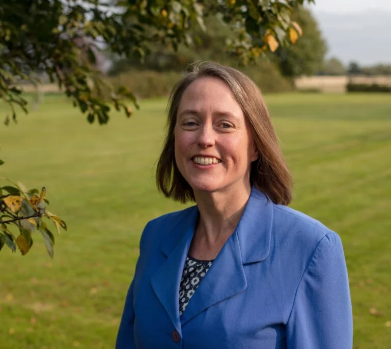 A smiling woman outdoors in a park with green grass and trees, wearing a blue jacket.