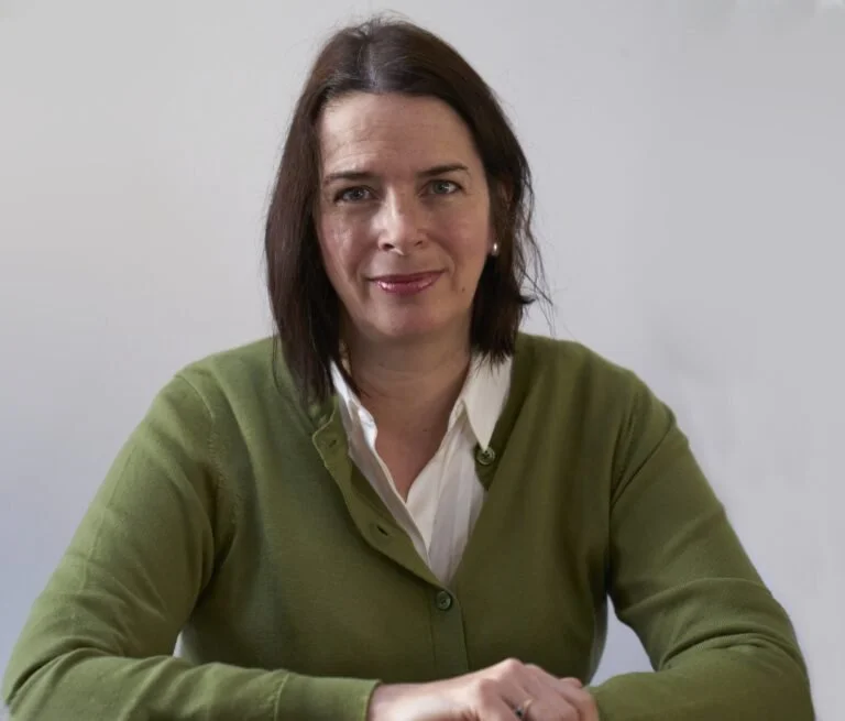 A woman with dark brown hair wearing a green cardigan over a white blouse, sitting at a table in front of a plain, light-colored background.