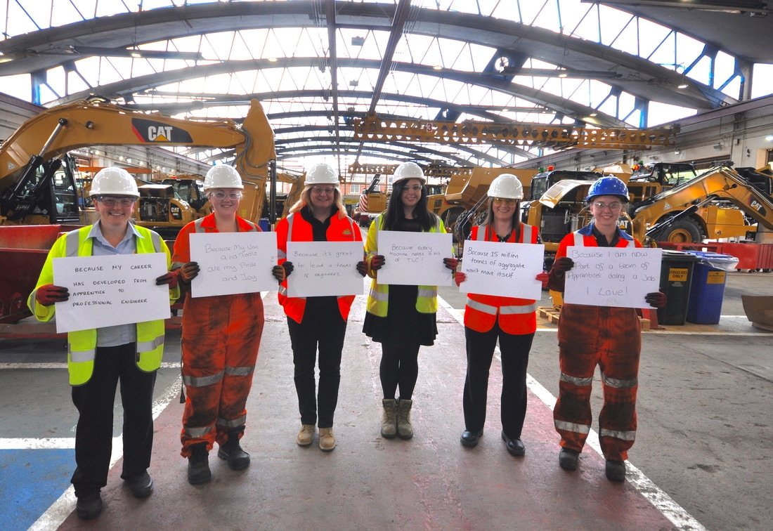 Six women in safety vests and hard hats standing inside a construction warehouse, holding whiteboards with messages. Large construction equipment, such as excavators, are visible in the background.