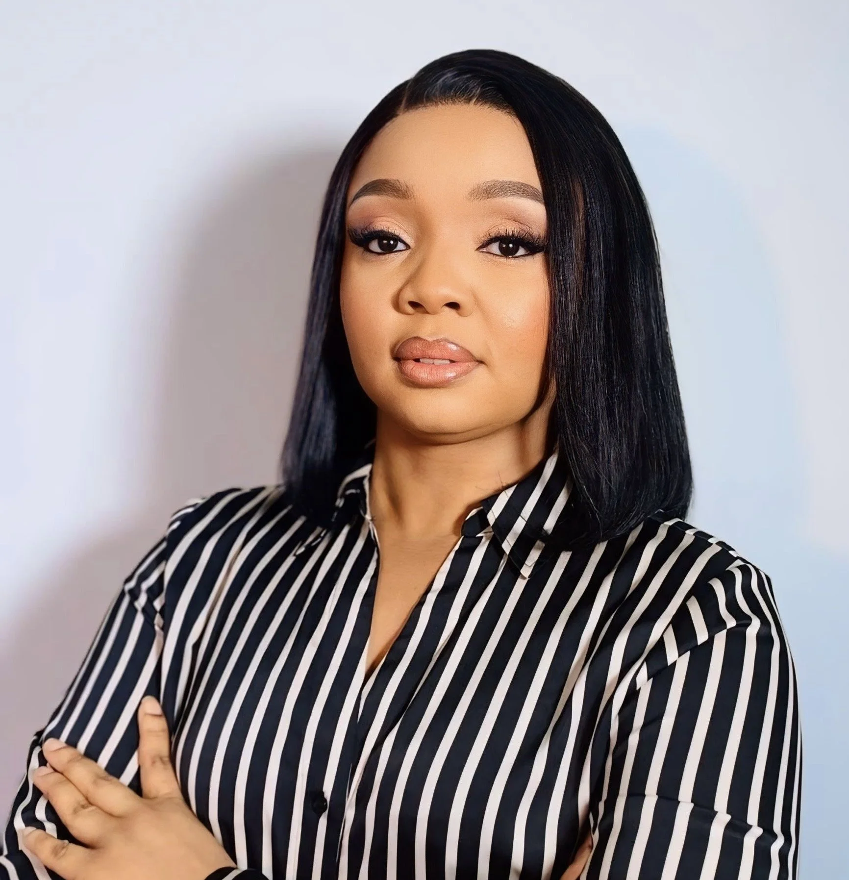 A woman with straight black hair and light makeup wearing a black and white striped shirt with rolled-up sleeves, standing with her arms crossed against a plain white background.
