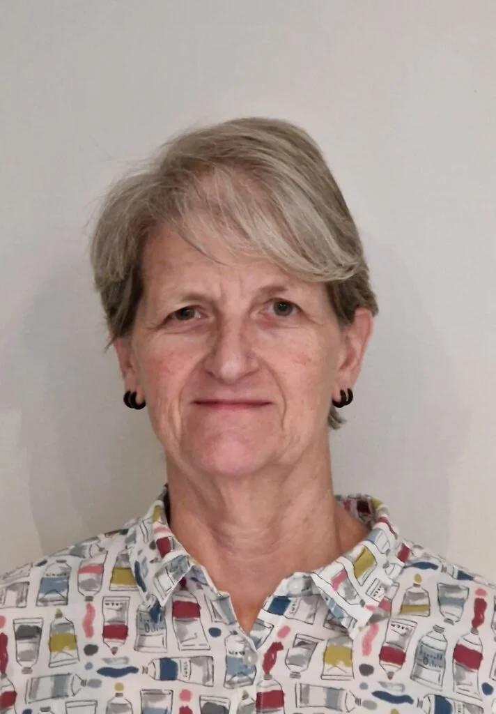 Close-up portrait of a middle-aged woman with short, light brown hair, wearing black earrings and a colorful patterned button-up shirt, standing against a plain light-colored background.