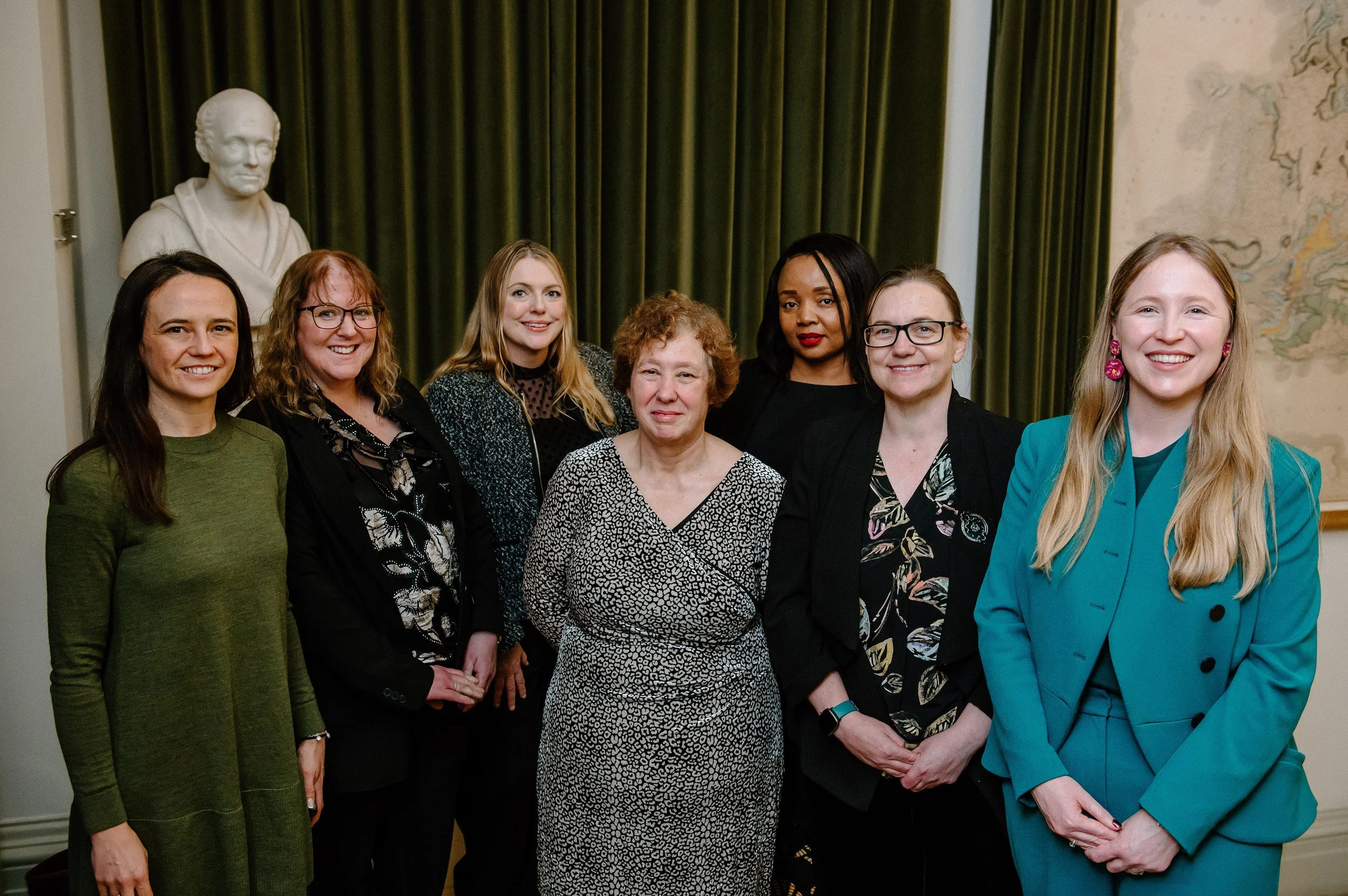 A group of seven women standing together indoors, with a bust sculpture and a world map in the background.