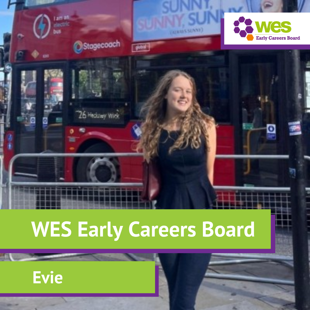A woman with light brown curly hair smiling outdoors, wearing a black top. Label says Board, displayed on a green banner.