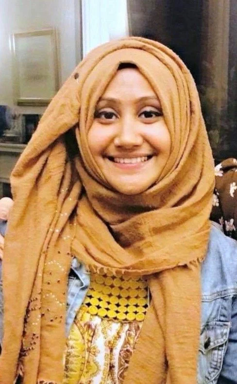 A woman wearing a tan hijab and a denim jacket, smiling at the camera in an indoor setting.