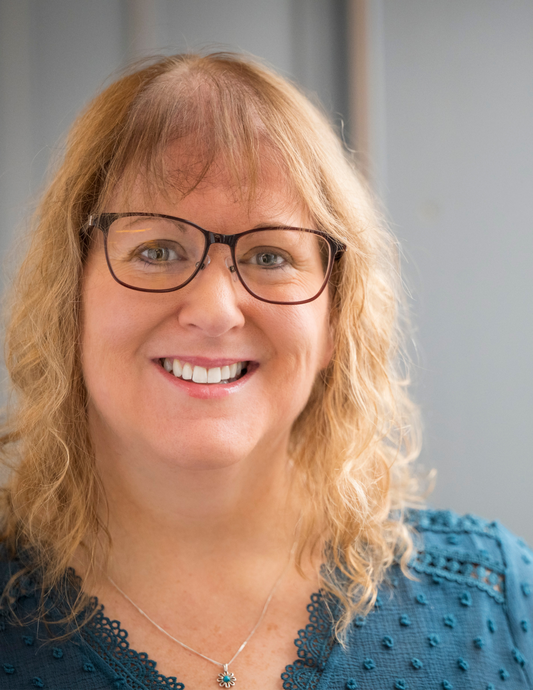 Close-up of a smiling woman with red curly hair and glasses, wearing a blue knitted top with lace details and a silver necklace with a flower pendant.