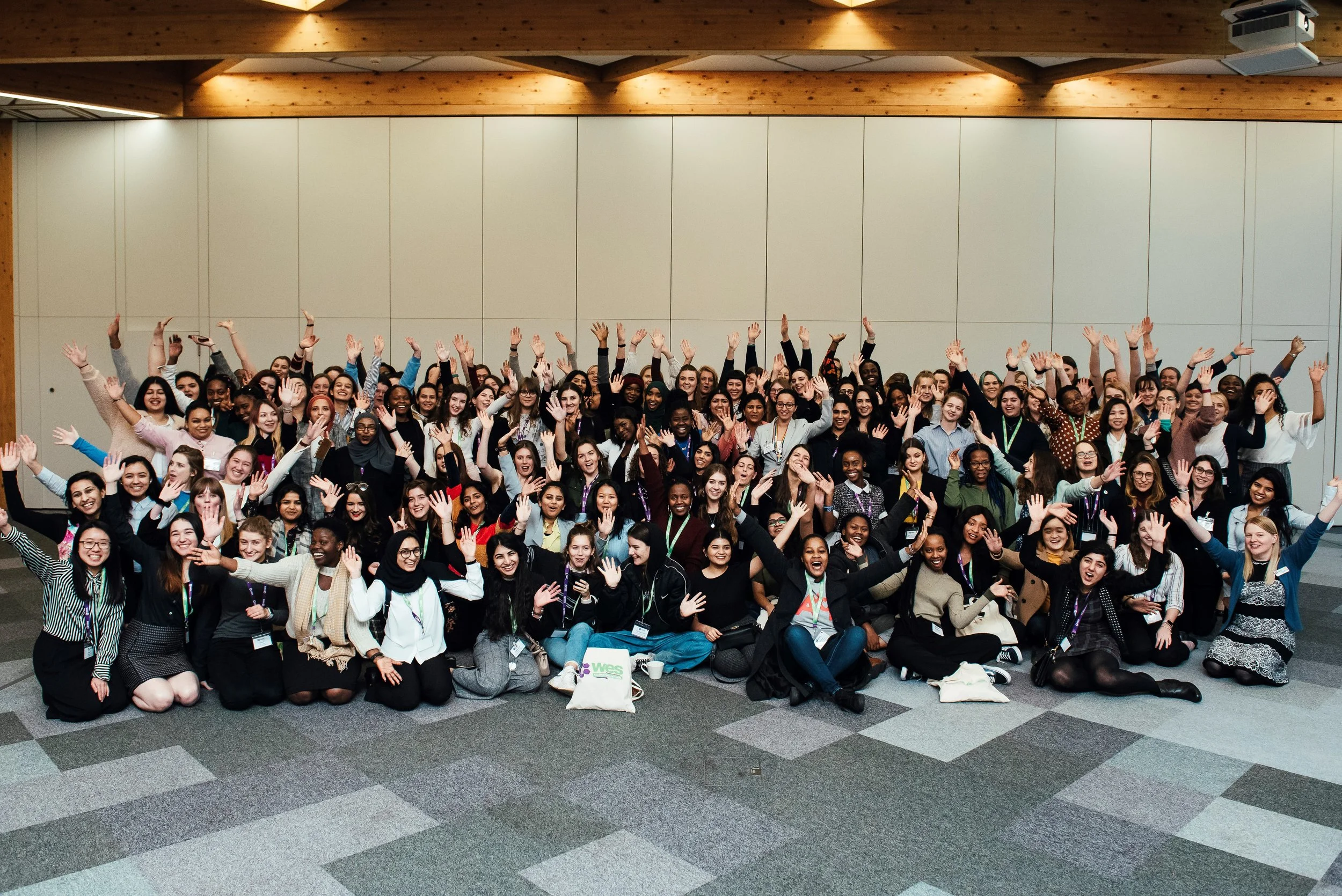 A large group of diverse people gathered together in a conference room, smiling and waving at the camera.