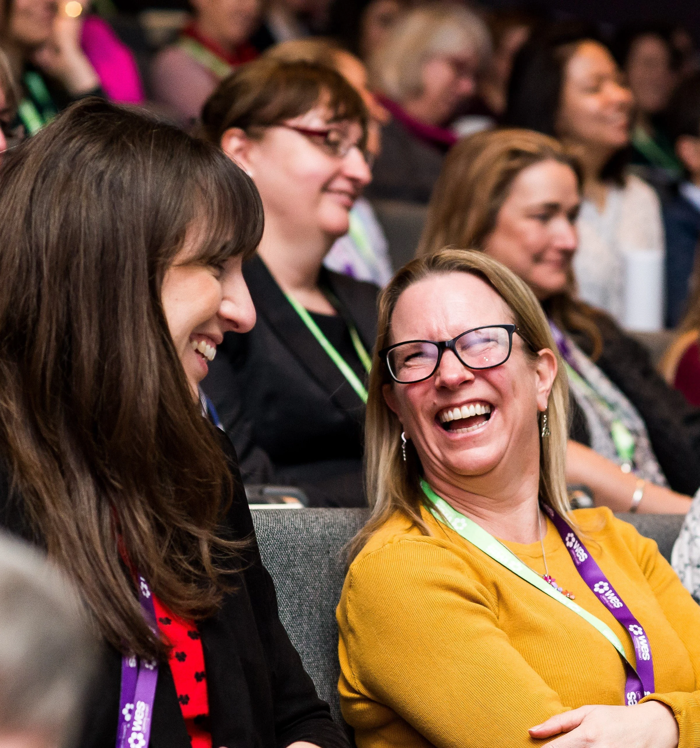 Group of people seated enjoying a moment of laughter, with some wearing glasses and smiling, at an indoor event.