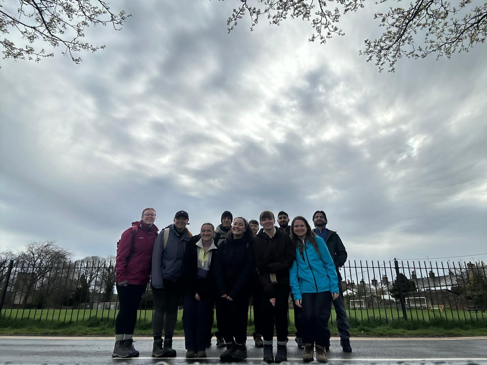 Group of nine smiling people standing outdoors on a sidewalk, with a metal fence and open grassy field behind them under a cloudy sky.