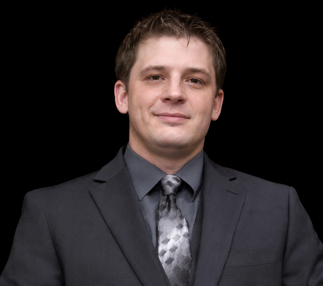 A smiling man in a dark suit, white shirt, and teal striped tie, with a boutonniere on his lapel, against a dark background.