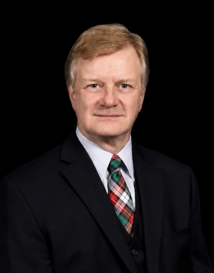A middle-aged man in a dark suit with a colorful plaid tie, posing against a black background.